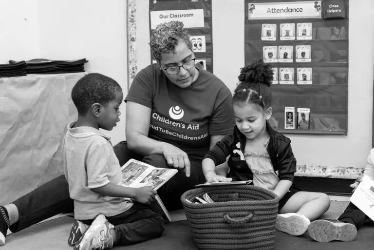 A teacher sits on a classroom floor with two young children, looking at picture books together.