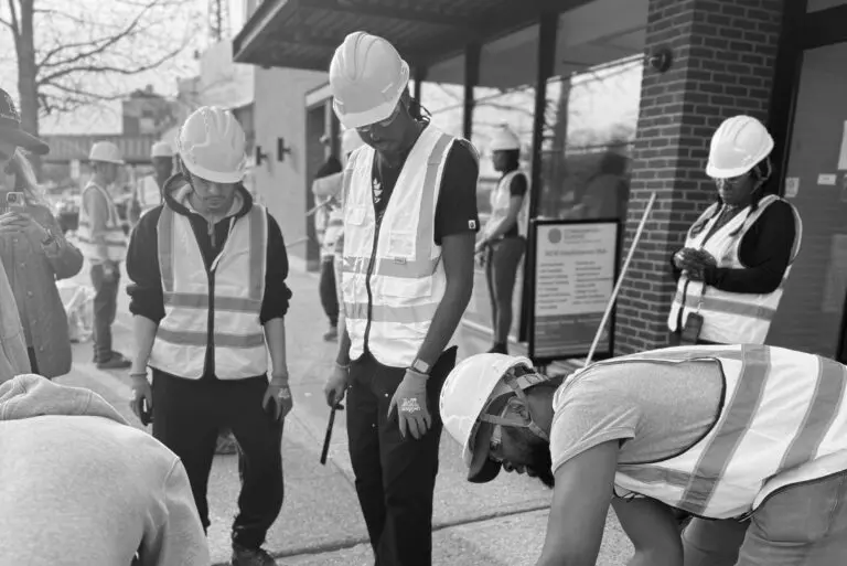 Participants in hard hats and reflective safety vests gather on a city sidewalk during a hands-on construction training session.